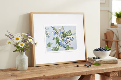 Framed blueberry block print on a wooden table with a vase of flowers and a bowl of berries.