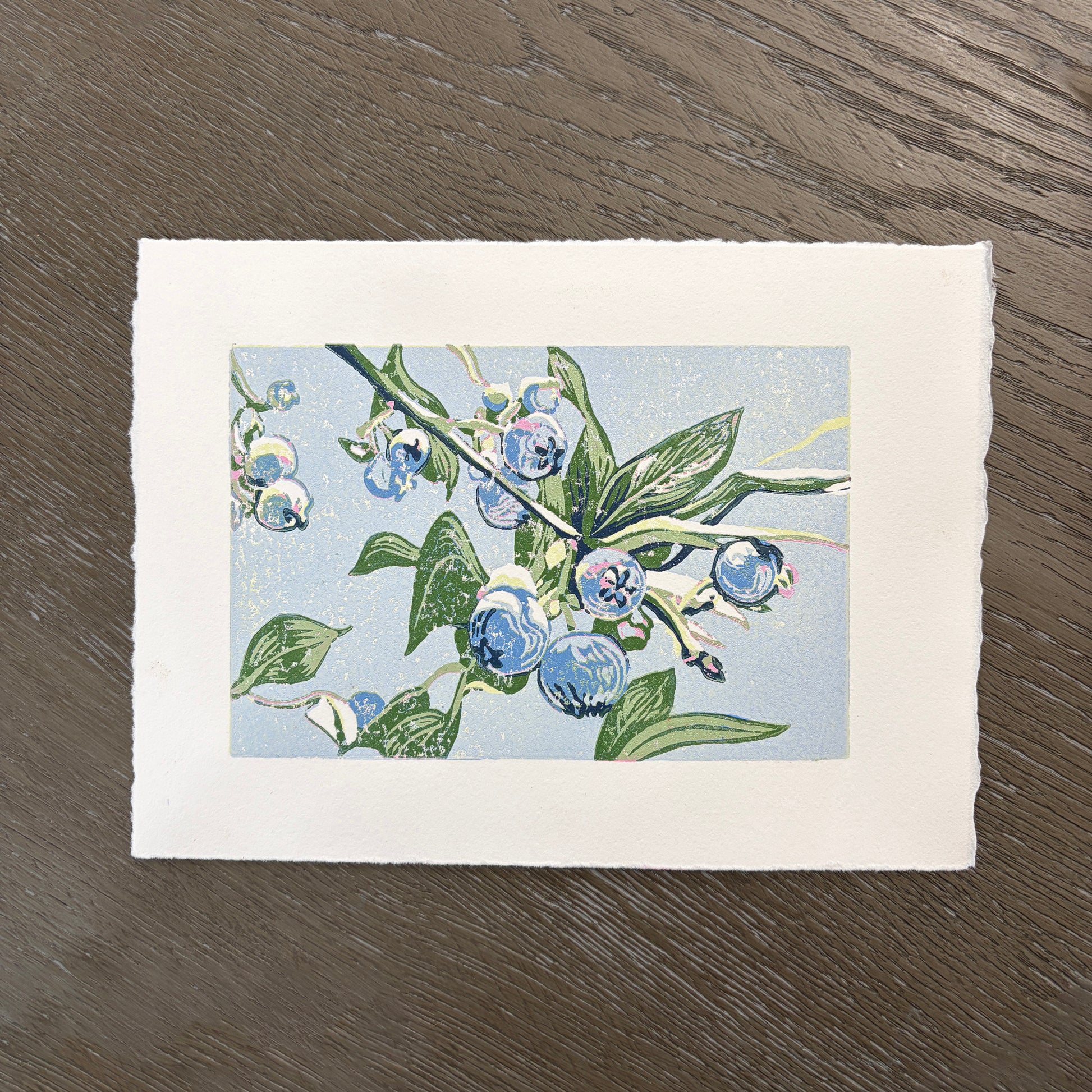 Deckled edge block print of blueberries and green leaves on a light blue background.