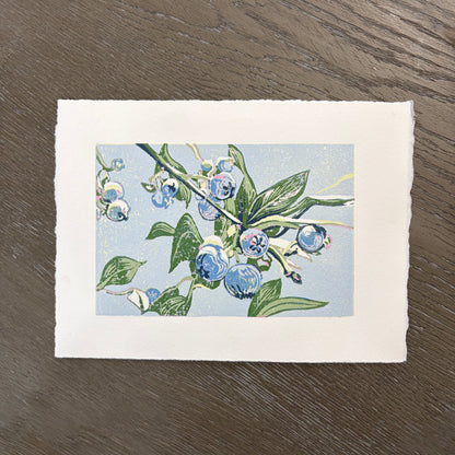 Deckled edge block print of blueberries and green leaves on a light blue background.