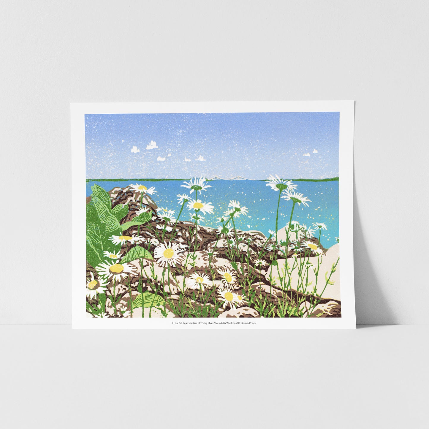 A fine art reproduction of "Daisy Shore," a 10" x 8" eight-color reduction block print of joyful daisies growing amongst limestone rocks on the Mackinac Island shore. Tucked back in the clear blue sky and turquoise water backdrop is the Mackinac Bridge.